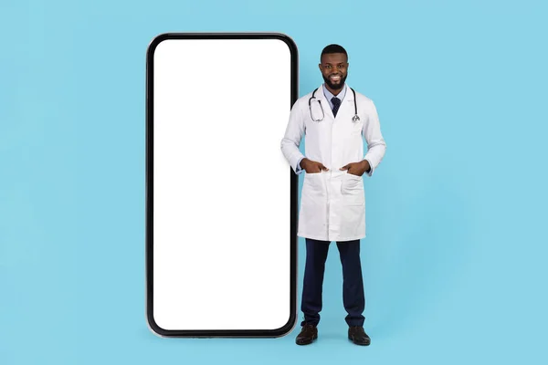 stock image Medical Offer. Smiling Black Doctor In Uniform Standing Near Huge Blank Smartphone Over Blue Background, Handsome Young African American Therapist Male Advertising Mobile App Or Website, Mockup