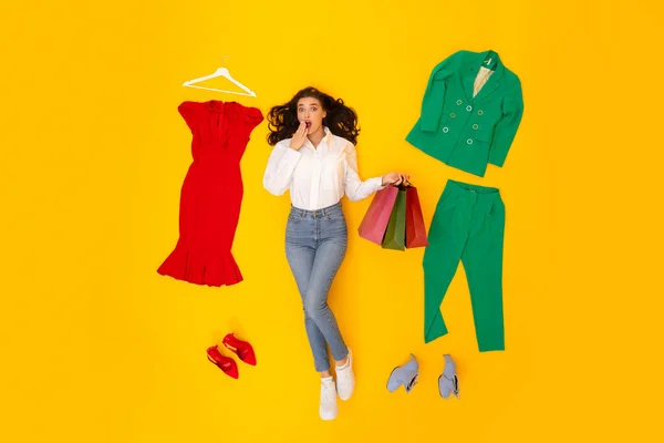 时尚与购物 女作家抱着购物袋躺在衣服和鞋子中间 看着相机 在黄色的工作室背景下在震惊中捂着嘴 以上视图拍摄 — 图库照片