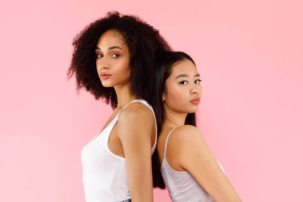stock image Portrait of young pretty asian and african american women standing back to back, posing together and looking at camera on pink background. Lifestyle diverse nationality people concept