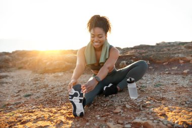 Spor giyimli genç bir kadın havluyla güneş doğarken okyanus sahilinde bir şişe suyla bacak esnetiyor. Vücut bakımı ve sağlık hizmetleri, pilates, sabah sporu, açık havada kilo verme.