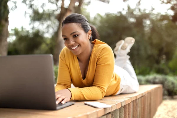 ラップトップで屋外で勉強している幸せなラテン語の女性学生 公園でベンチに横たわっている間 女性のフリーランサーは コンピュータ上で作業します 画面を見て — ストック写真