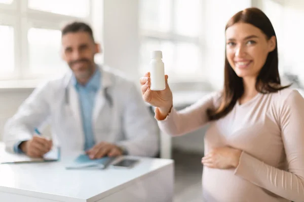 产前护理概念 快乐的年轻孕妇 手里拿着一罐药丸或维生素 坐在诊所里与男产科医生会诊 有选择性地集中精神 — 图库照片