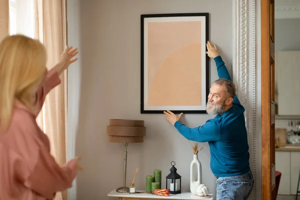 陽気なシニア配偶者壁に飾られた現代のリビングルームに一緒に改装後の家に立ってポスターをぶら下げて インテリアデザインのコンセプト 夫への選択的焦点 — ストック写真
