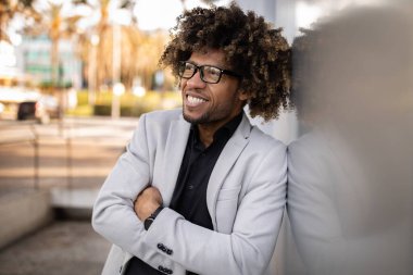 Heyecanlı Afro-Amerikan orta yaşlı iş adamı takım elbiseli çapraz kollarla poz veriyor, duvara yaslanıyor ve başka tarafa bakıyor başarılı bir iş toplantısından sonra mola veriyor, dışarıda duruyor.
