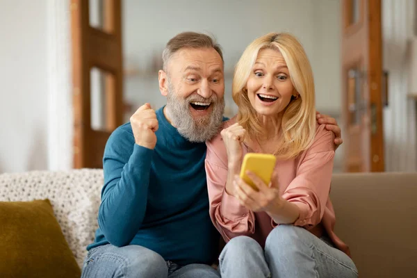 stock image Joyful Senior Couple Using Smartphone Reading Message And Shaking Fists Celebrating Big Luck Sitting On Couch At Home. Wow Mobile Offer And Application Advertisement Concept