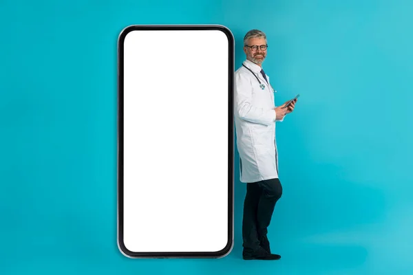 快乐的微笑英俊的白发苍苍的中年男人医生站在大手机旁边 白白的屏幕 拿着手机 与病人在线聊天 蓝色背景 — 图库照片