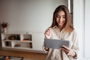 Akıllı evi kontrol etmek için günlük kullanım tabletinde gülümseyen güzel Asyalı genç bayan, çalışmanın ve aydınlık oturma odasının içinde çalışmanın keyfini çıkar. Teknoloji ile iş, sosyal ağlarda sohbet