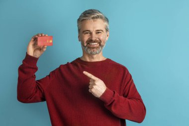 Neşeli, yakışıklı, gri saçlı, olgun bir adam kırmızı plastik banka kartına bakıyor ve gülümsüyor. Temassız ödeme öneriyor. Kolay hızlı bankacılık, mavi stüdyo geçmişi, kopyalama alanı.