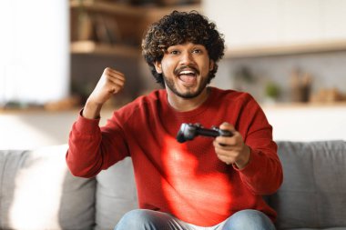 Duygusal Hintli adam evde joystick ile video oyunu oynuyor, neşeli genç doğu erkeği yumruğunu sallıyor ve oturma odasında kanepede otururken zaferi kutluyor, milenyum adamı oyun oynamaktan zevk alıyor.