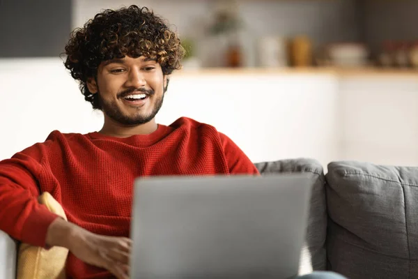 Online Communication. Happy Young Indian Man Using Laptop While Sitting On Couch At Home, Smiling Handsome Eastern Guy Typing On Computer And Relaxing On Sofa In Living Room, Closeup Shot