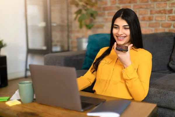 スペイン人女性がノートパソコンを使ってデスクに座っているのを笑顔にしたり 電話で話したり 音声メッセージを指示したり オンライン翻訳アプリや音声認識ソフトウェアを使用したり バーチャルアシスタント — ストック写真