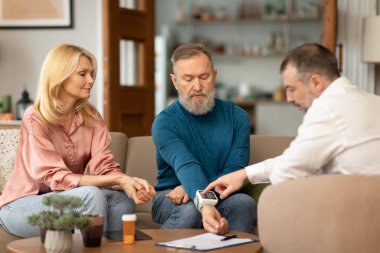 Erkek doktor, yetişkin bir erkek için bileğinde tıbbi aletle kan basıncını ölçerken yaşlı çift evde oturup sağlıkla ilgileniyordu. Seçici Odaklanma