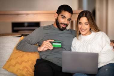 Bin yıllık Arap kocası beyaz karısına sarılıyor, dizüstü bilgisayar ve kredi kartı kullanıyor, oturma odasında online alışveriş yapıyor. İndirim, sipariş, cihaz, evde nakit para.
