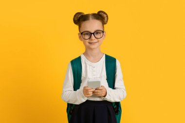 Elinde akıllı telefon ve sırt çantasıyla sarı stüdyoda poz veren, cep telefonu kullanan ve kameraya gülümseyen küçük kız öğrencinin portresi.