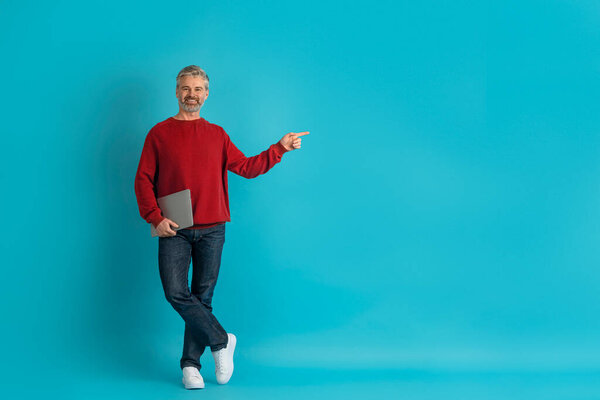 Smiling attractive grey-haired middle aged man wearing casual outfit with laptop in his hand posing on blue studio background, showing empty space for advertisement, recommending nice website