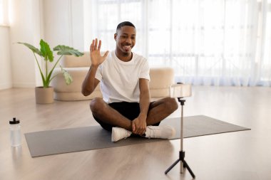 Spor kıyafetli neşeli, yakışıklı siyah adam yoga minderinde oturuyor, cep telefonu kamerasına el sallıyor ve gülümsüyor, evden yayın yapan erkek fitness blogcusu, kopyalama alanı. Fitness günlük yazma kavramı