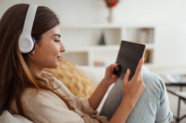 Kablosuz kulaklıklı olumlu genç Avrupalı kadın tabletten video izliyor, oda içi, profil, yakın çekim yapıyor. Sosyal ağlarda sohbet, çevrimiçi alışveriş, müzik ve ders uygulaması