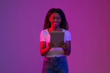 Gülümseyen Siyah Kadın, Neon Light 'ın içinde dikilirken dijital tablet kullanıyor, Neşeli Afro-Amerikalı Kadın, Mor Stüdyo Arkaplanı Üzerine Poz Verirken, İnternet' i inceliyor, Boşluğu Kopyala