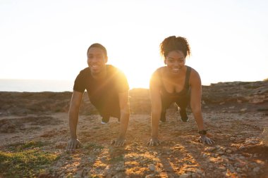 Gülümseyen binlerce yıllık Afro-Amerikan kadın ve spor giyimli adam güneş doğarken okyanus sahilinde şınav çekiyorlar. Birlikte kaslar, vücut bakımı ve sağlık hizmetleri için sabah egzersizi, açık havada kilo kaybı.