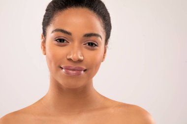 Güzel, üstsüz, kameraya gülümseyen, gri stüdyo arka planında poz veren, parlak cildini, fotokopi alanını ve afişini gösteren siyah bir kadının yakın çekimi. Kozmetoloji kavramı