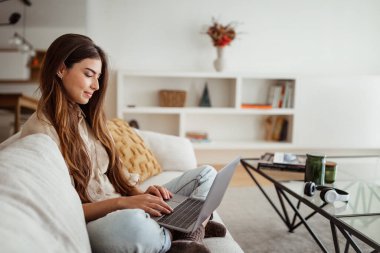 Minimalist oturma odasında bilgisayarda yazı yazan neşeli, Avrupalı bir kadın var. Çalışma, serbest çalışma, iş ve iş uzaktan evde, cihaz ve yeni normal