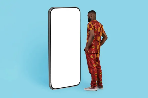 移动报价 传统服饰中的非洲年轻人站在蓝色背景下 用白色屏风看大块空白智能手机 黑人查新的应用 大学生活 — 图库照片