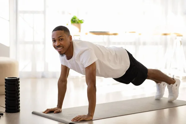 穿着运动服 穿着运动服的英俊黑人年轻人躺在瑜伽垫上 在家里做早操 使用健身工具 看着相机笑着 复制空间 全身而退 — 图库照片