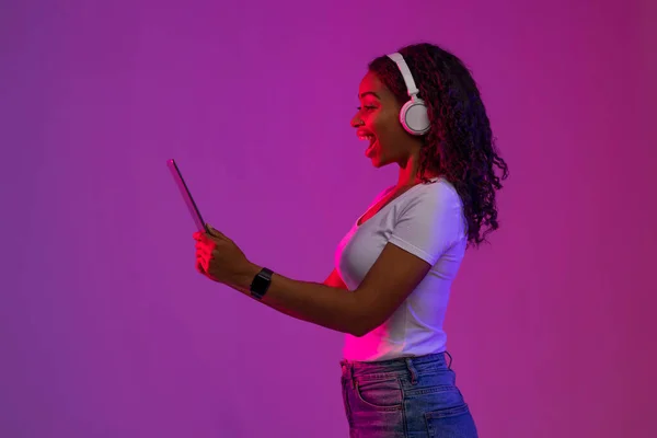 Excited Black Woman Playing Video Games On Digital Tablet In Neon Light, Happy Young African American Lady Wearing Wireless Headphones Using Modern Gadget While Standing Over Purple Background