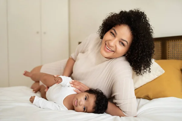 Glad young african american lady plus size and little child lie on bed, enjoy baby care, relax together, play in bedroom interior. Motherhood, love, relationship mom and kid at home