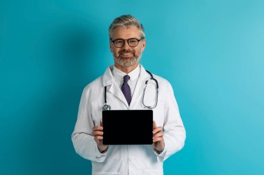 Neşeli, yakışıklı, orta yaşlı beyaz doktor siyah ekranlı dijital tablet gösteriyor, güzel bir tele sağlık web sitesi veya uygulama öneriyor, mavi stüdyo geçmişi, maket, kopyalama alanı öneriyor.
