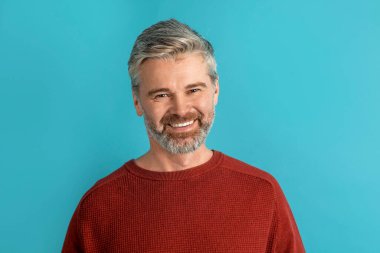 Neşeli, gülümseyen, beyaz saçlı, gri sakallı orta yaşlı bir adamın portresi. Kameraya gülümsüyor, mavi stüdyo arka planında tek başına poz veriyor.