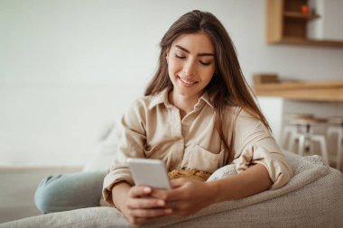 İnternette sörf yapmak için bir cihaz. Minimalist oturma odasında telefonla yazan neşeli Avrupalı bayan, fotokopi alanı. Sosyal medya sohbeti, evde uygulama, iyi haber.