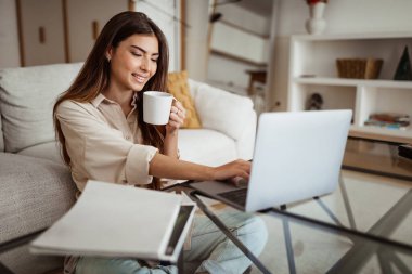 Binlerce yıllık mutlu Avrupalı kadın bilgisayarda yazı yazıyor, minimalist oturma odasında kahve içiyor. Serbest meslek, iş ve iş uzaktan evde, aletle öğrenim