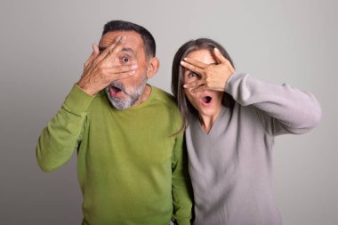 Shocked frightened old european man and woman with open mouth covers eyes with hands on gray studio background, ad and offer. Fear, scare, people emotions and facial expression clipart