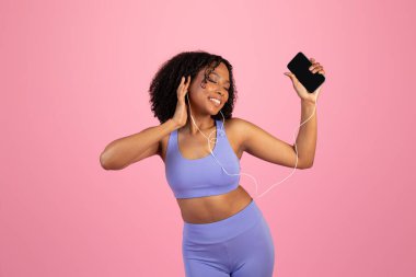 Mutlu Milenyum Afro-Amerikan kadın spor giyim, kulaklıklar boş ekranlı telefon kullanır, müzik dinler, pembe stüdyo arka planında dans eder. Ses uygulaması, eğlence, spor, vücut bakımı