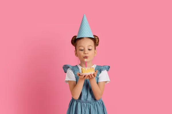 誕生日ケーキにパーティーの帽子を吹いてキャンドルを身に着けているかわいい女の子 甘いB日の御馳走の幸せな十代の女性の子供を保持します ピンクのスタジオの背景に立っている間願いを作ります スペースをコピー — ストック写真