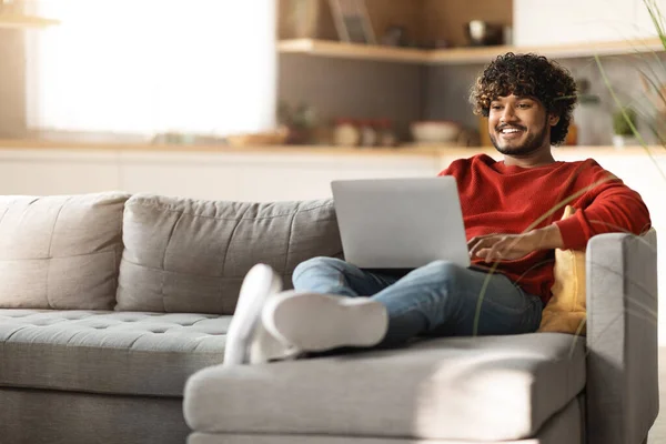 Freelance Concept. Handsome Young Indian Man Using Laptop While Sitting On Couch At Home, Millennial Eastern Guy Working Remotely On Computer In Living Room, Enjoying Online Work, Copy Space