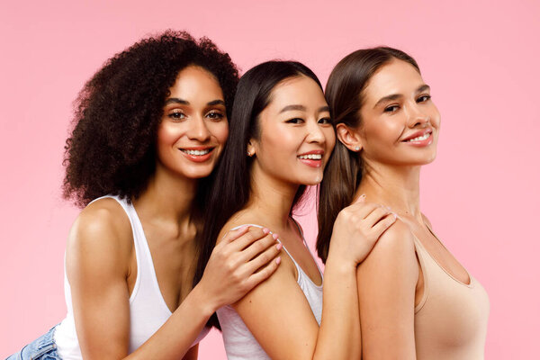 Young pretty asian, caucasian and black women posing together and smiling at camera, standing on pink background. Lifestyle diverse nationality people concept