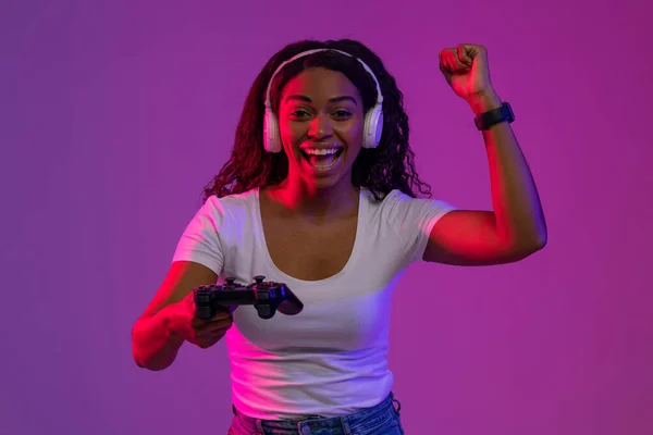 Video Gaming Concept. Euphoric African American Woman With Joystick Celebrating Game Win, Cheerful Black Lady Raising Fist And Exclaiming With Excitement, Standing In Neon Light On Purple Background