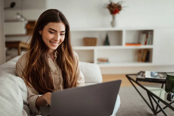 Smiling millennial european female typing on computer, has chat and video call in minimalist living room interior. Gadget for study, freelance, work and business at home remotely