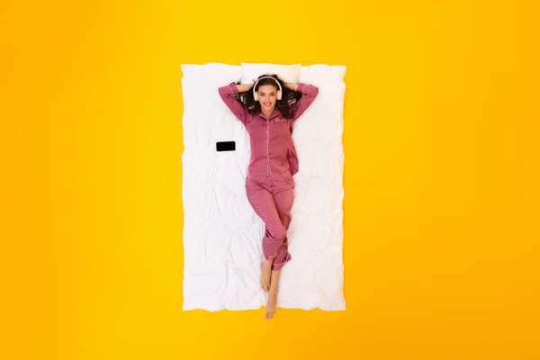 快乐的女士带着耳机在网上听音乐在黄色工作室背景下躺在床上 微笑在相机前 音乐移动应用概念 — 图库照片
