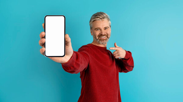 Happy handsome grey-haired middle aged man in casual showing great mobile app or online offer, holding cell phone with white empty screen in his hand, blue background, banner, mockup