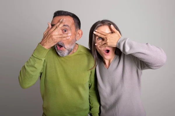 Shocked frightened old european man and woman with open mouth covers eyes with hands on gray studio background, ad and offer. Fear, scare, people emotions and facial expression