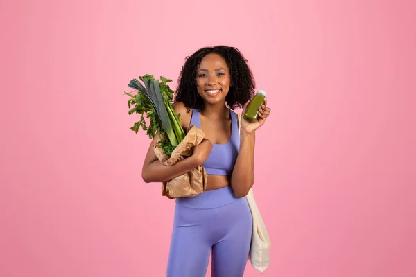 スポーツウェアショーの紙袋に緑の野菜とピンクのスタジオの背景に隔離されたスムージーボトルで千年黒の女性を笑顔 体重減少 ボディ ヘルスケア ベジタリアンフード — ストック写真