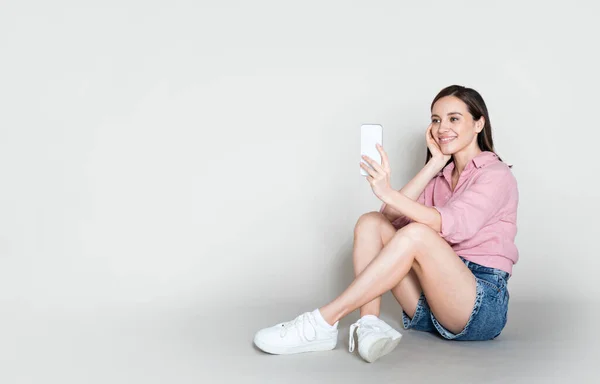 喜びに満ちた幸せな美しいブルネット若い女性が夏服を着て床に座って 携帯電話で自撮り グレーのスタジオの背景 広告のためのコピースペースを持つパノラマ — ストック写真