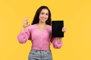 Bin yıllık Arap kadın günlük gösteri tabletinde boş ekranlı, tamam sarı arka planda el izole edilmiş bir tabela, stüdyo. Tavsiye, aygıt sunumu, tavsiye web sitesi ve uygulama