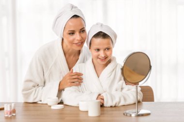 Bornoz ve havlu giymiş güzel bir anne ve kız birlikte yüz kremi sürüyorlar, mutlu anne ve tatlı küçük kız çocuk makyaj masasında oturuyor ve aynaya bakıyor, güzellik rutininin keyfini çıkarıyorlar.