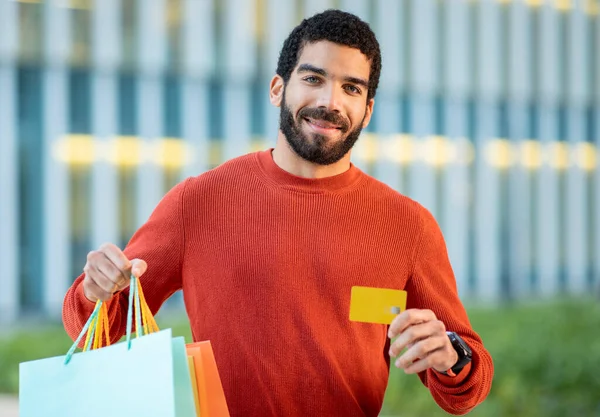 Happy Middle Eastern Man Shopping Paying With Credit Card, Posing Showing Paper Shopper Bags To Camera Standing Outdoors Near Mall. Great Sales And Bank Offer. Selective Focus