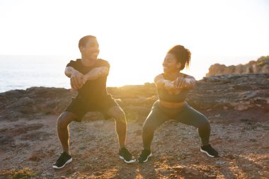 Genç Afro-Amerikan kız arkadaşı ve spor giyimli bir adam okyanus sahilinde çömelip, güneş doğarken sabah sporunun tadını çıkardıkları için mutluyum. Vücut bakımı ve sağlık hizmetleri birlikte, dışarıda kilo kaybı.