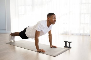 Motive olmuş yakışıklı, Afro-Amerikan sporcu, spor kıyafetleriyle evde spor yapıyor, spor paspası üzerinde duruyor, dambıllarla eğiliyor, fotokopi odasına bakıyor. Spor, spor, spor.